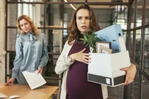 A pregnant woman carrying a box of stuff out of an office while another coworker is smiling in the back.