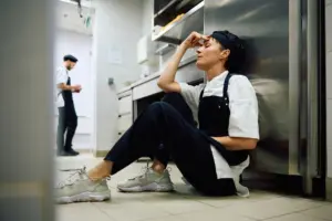 A tired chef sitting on the floor of a kitchen.