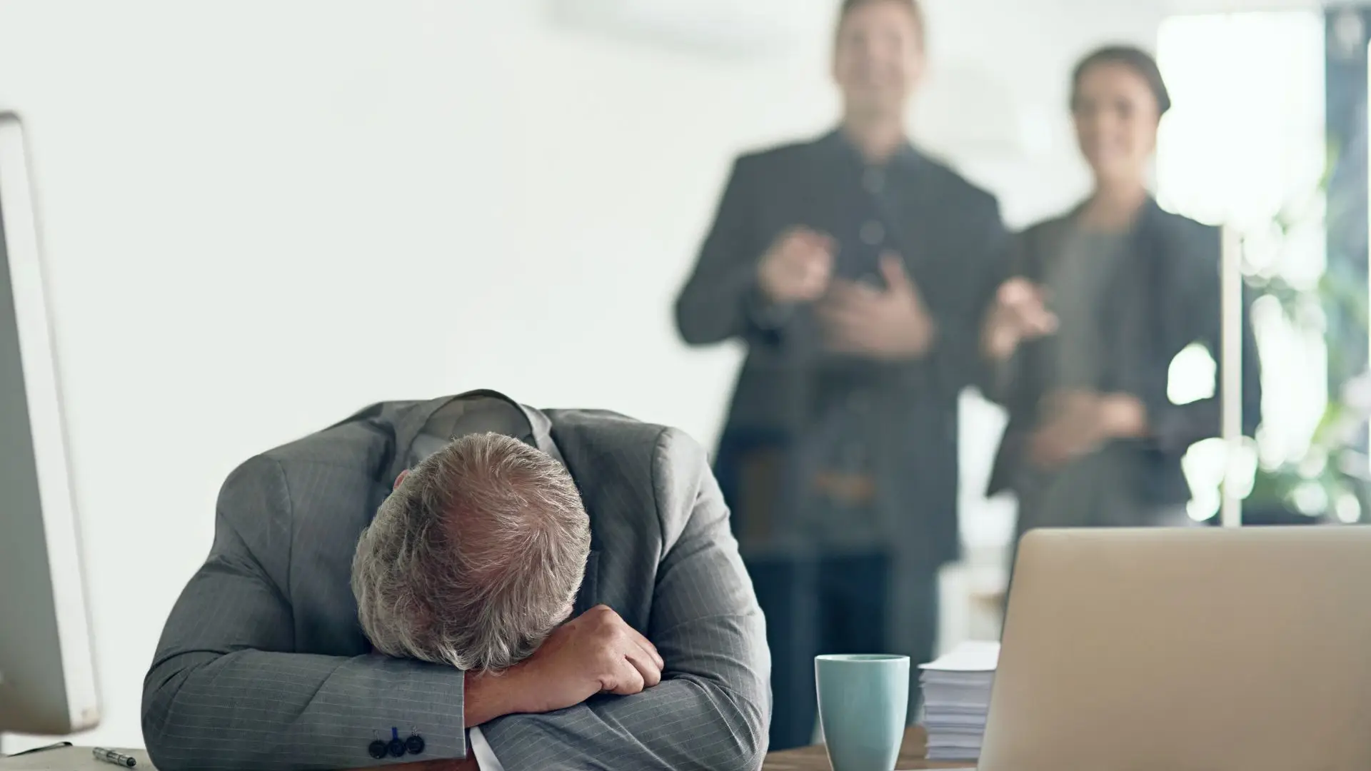 An older worker putting his head down in misery, while two coworkers are mocking him in the background.