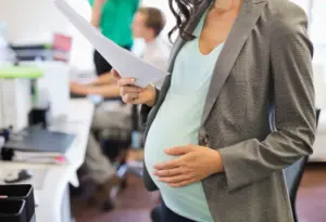 A pregnant worker holding onto her belly while looking at paperwork.
